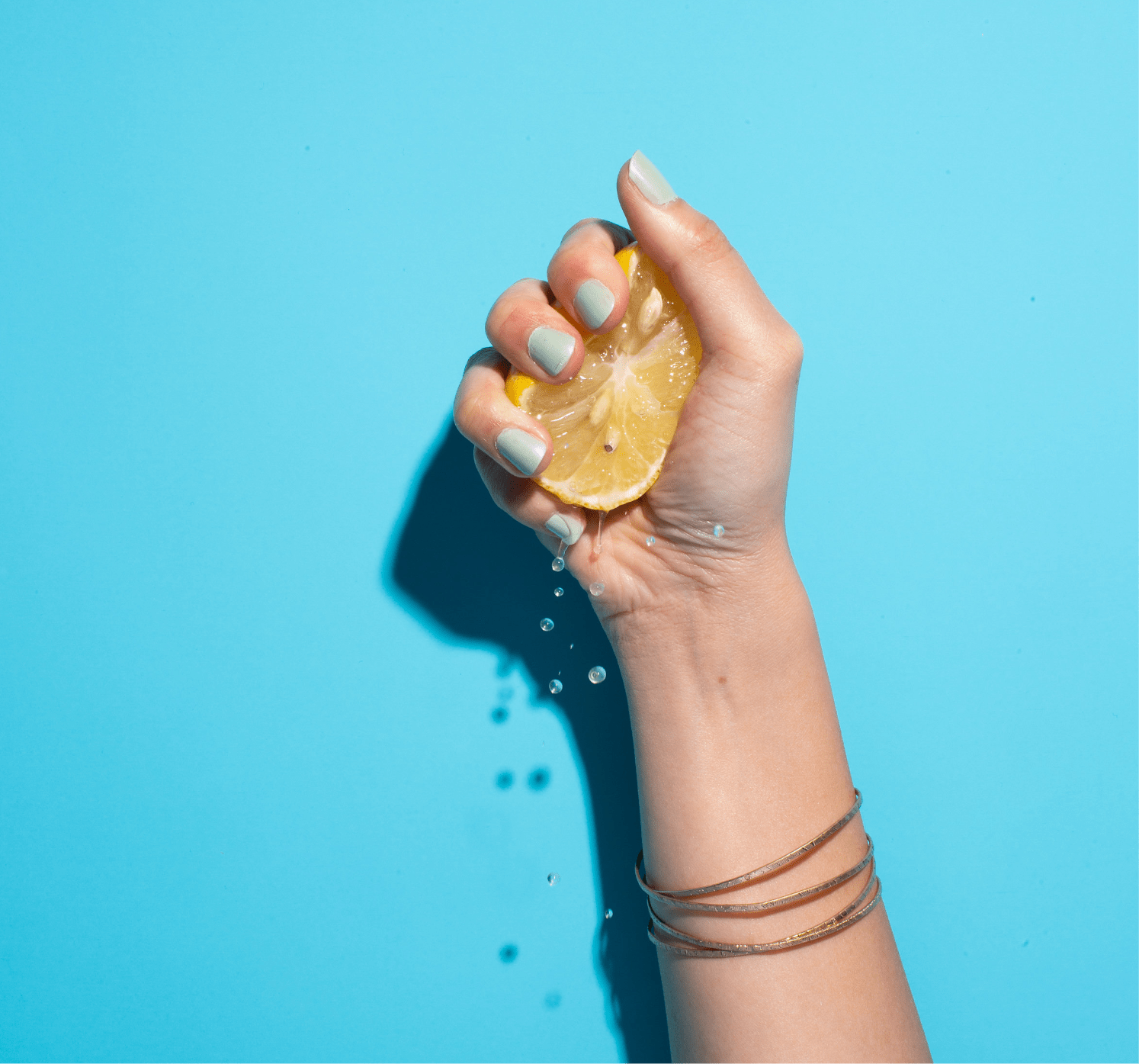 A hand with mint green nails squeezes a lemon against a vibrant blue background, releasing zesty juices and sparkling water droplets.