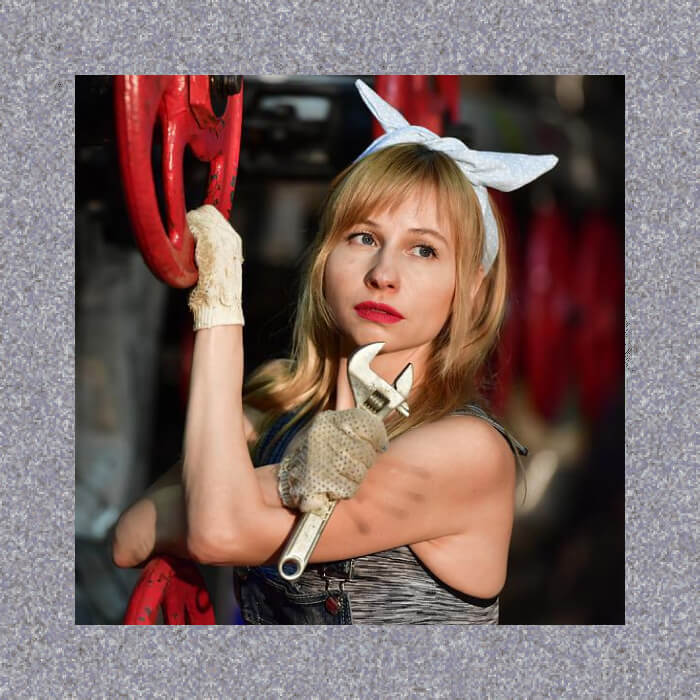 A woman in a bandana and gloves skillfully holds a wrench, poised confidently beside red machinery wheels, embodying the essence of mechanical prowess.