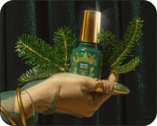 Hand holding a green Poo~Pourri Funk Fir toilet spray bottle with a gold cap between two pine branches against a dark green velvet background