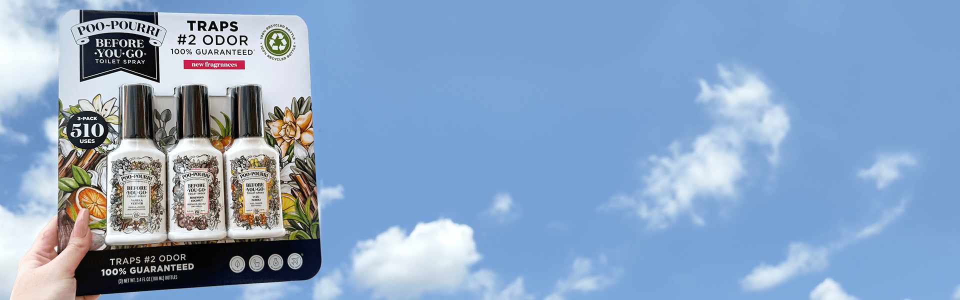 Hand holding a 3 pack of Poo-Pourri toilet sprays against a bright blue sky with scattered white clouds.
