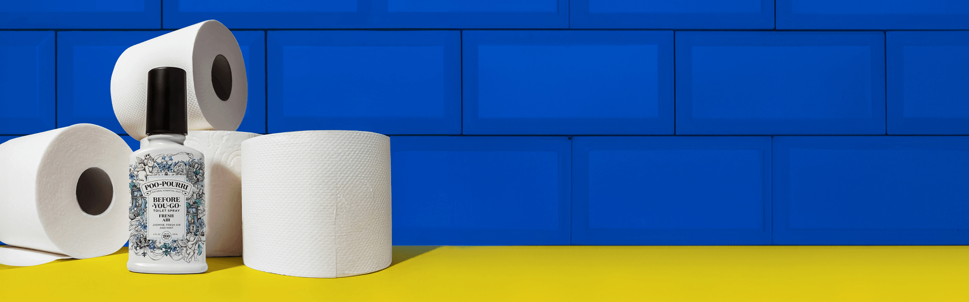 A bottle of Poo-Pourri “Before You Go” toilet spray in the Fresh Air scent is displayed among several white toilet paper rolls on a bright yellow surface. The background features a vivid blue tiled wall, creating a bold and contrasting color scheme.