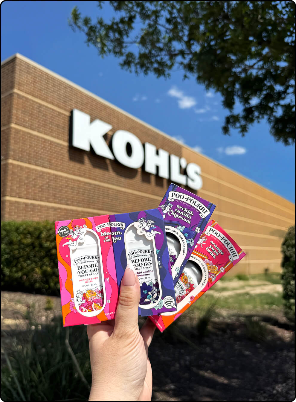 Hand holding colorful Poo~Pourri bottles and pocket sprayers in the Orange Yuzy Twist and Orchid Vanilla Blossom scents in front of a Kohl's store.