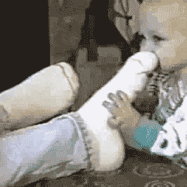 A baby giggles playfully as they nibble on a sock-covered foot, grasping it with both tiny hands.