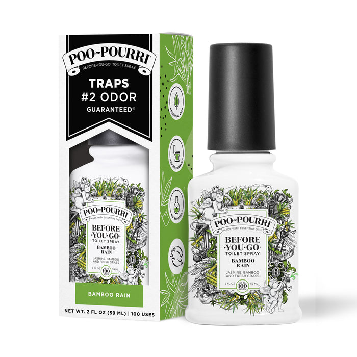 A ~Poo-Pourri “Before You Go” toilet spray in the Bamboo Rain scent is displayed alongside its packaging against a white background. The bottle features an ornate label with cherubs, toilet paper, and bamboo elements. The packaging highlights key product benefits, including its ability to trap odors, the use of essential oils, and the absence of aerosols. The scent is described as a blend of jasmine, bamboo, and fresh grass. The bottle contains 2 fl oz (59 ml) for up to 100 uses.