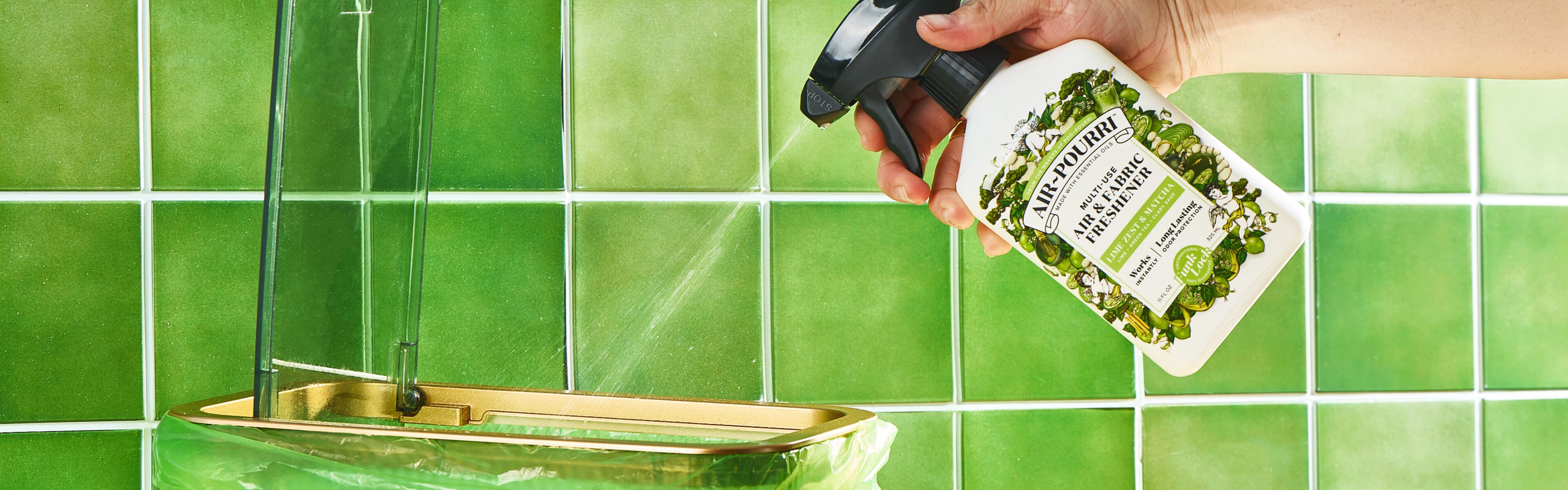 A hand holding a spray bottle of Air~Pourri Lime Zest and Matcha, spraying a fine mist onto a glass and gold sink against a green tiled wall.