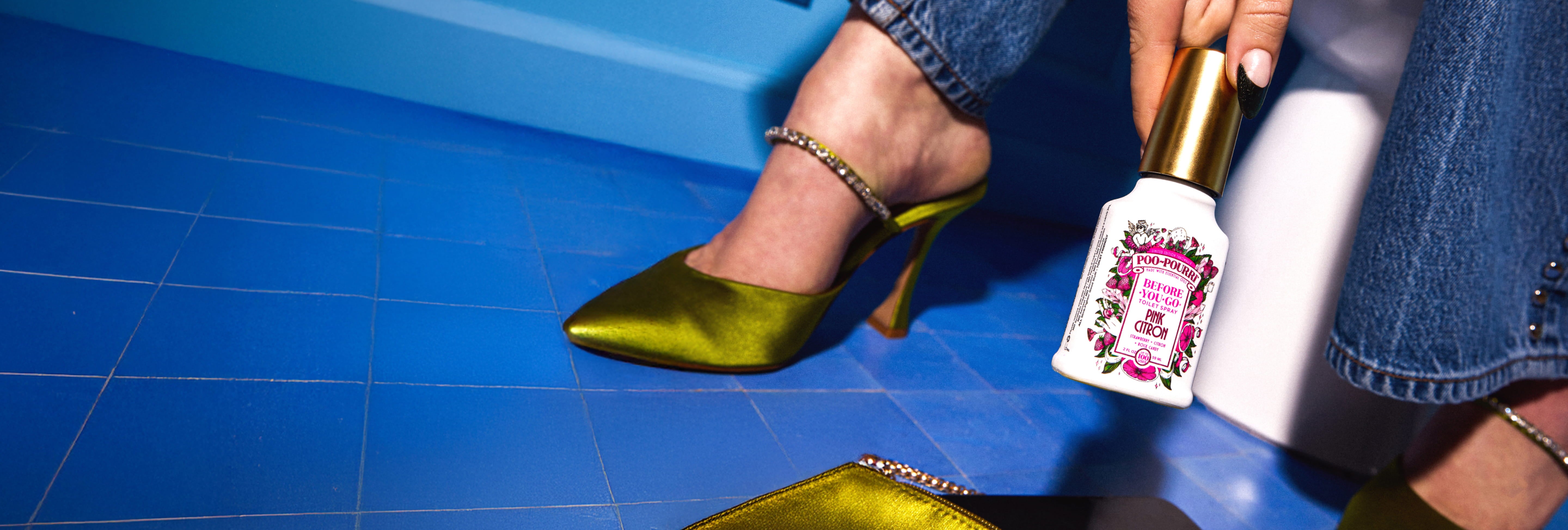 Person wearing green high heels with a blue floor holding a Poo~Pourri Pink Citron bottle in the foreground