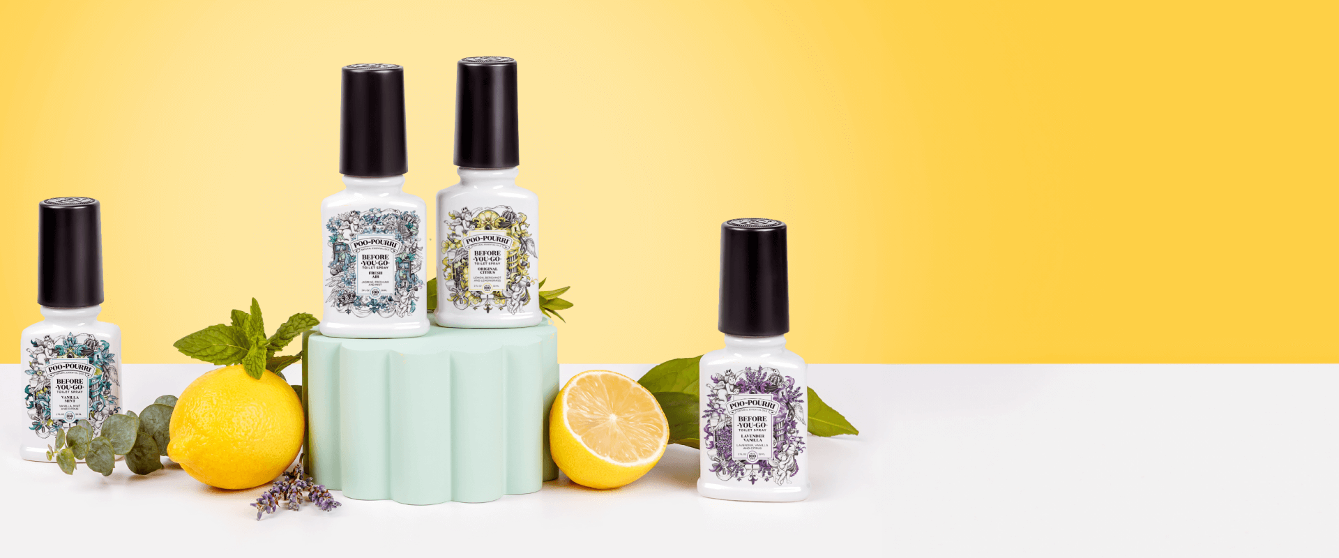 A display of four Poo-Pourri “Before-You-Go” toilet sprays in white bottles with black caps, arranged on a white and pastel green surface with a bright yellow background. Each bottle features ornate labels with different floral designs. The setup is accented with fresh lemons, lemon halves, mint leaves, lavender, and eucalyptus.