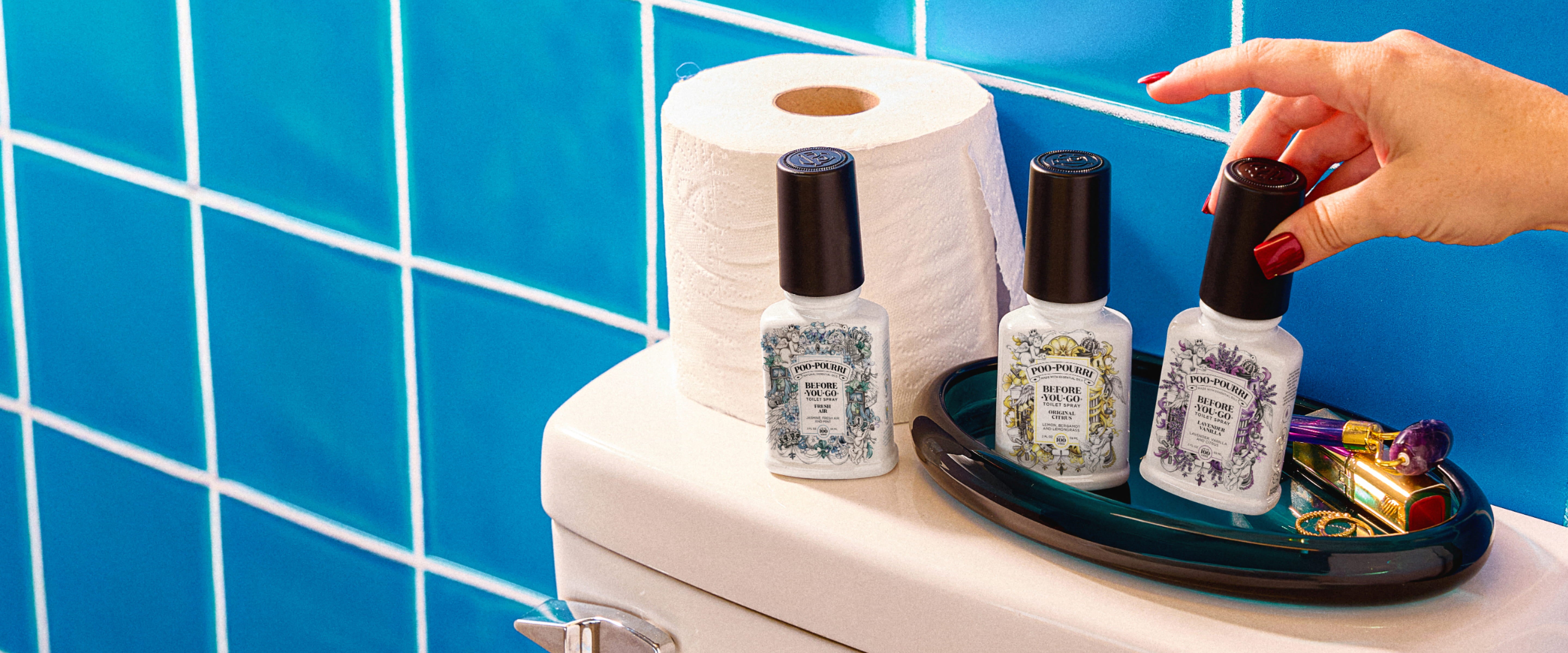Poo~Pourri bottles on a tray with toilet paper against a blue tiled bathroom wall.
