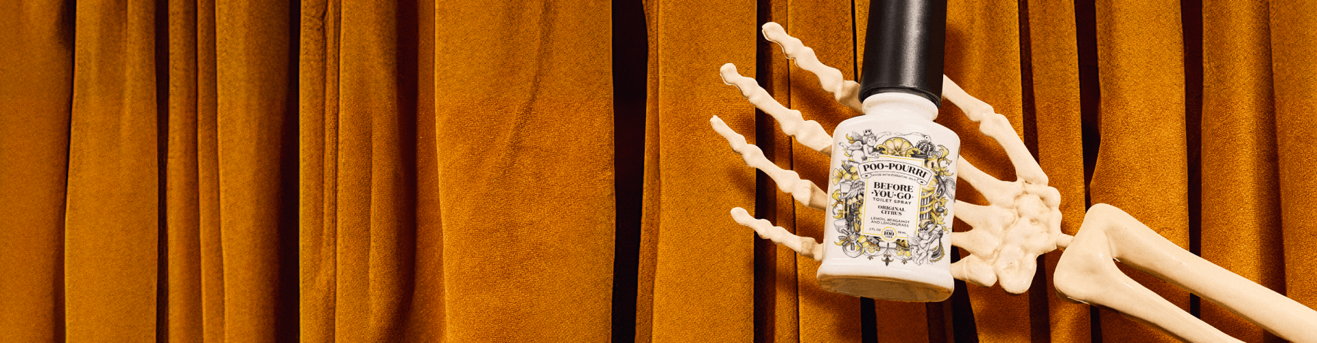 Bottle of Poo~Pourri Original Citrus toilet spray held by a skeleton hand against an orange curtain background