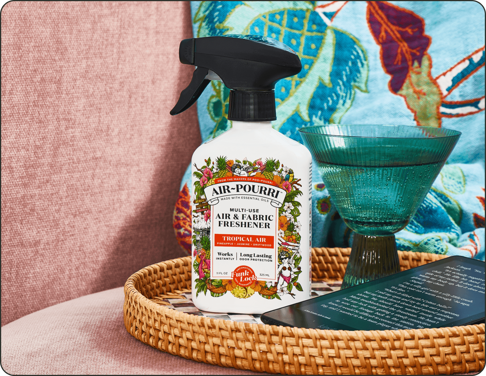 A bottle of Air-Pourri Tropical Air sits on a woven tray beside a teal glass and a book, set against a pink upholstered chair and a colorful patterned textile.