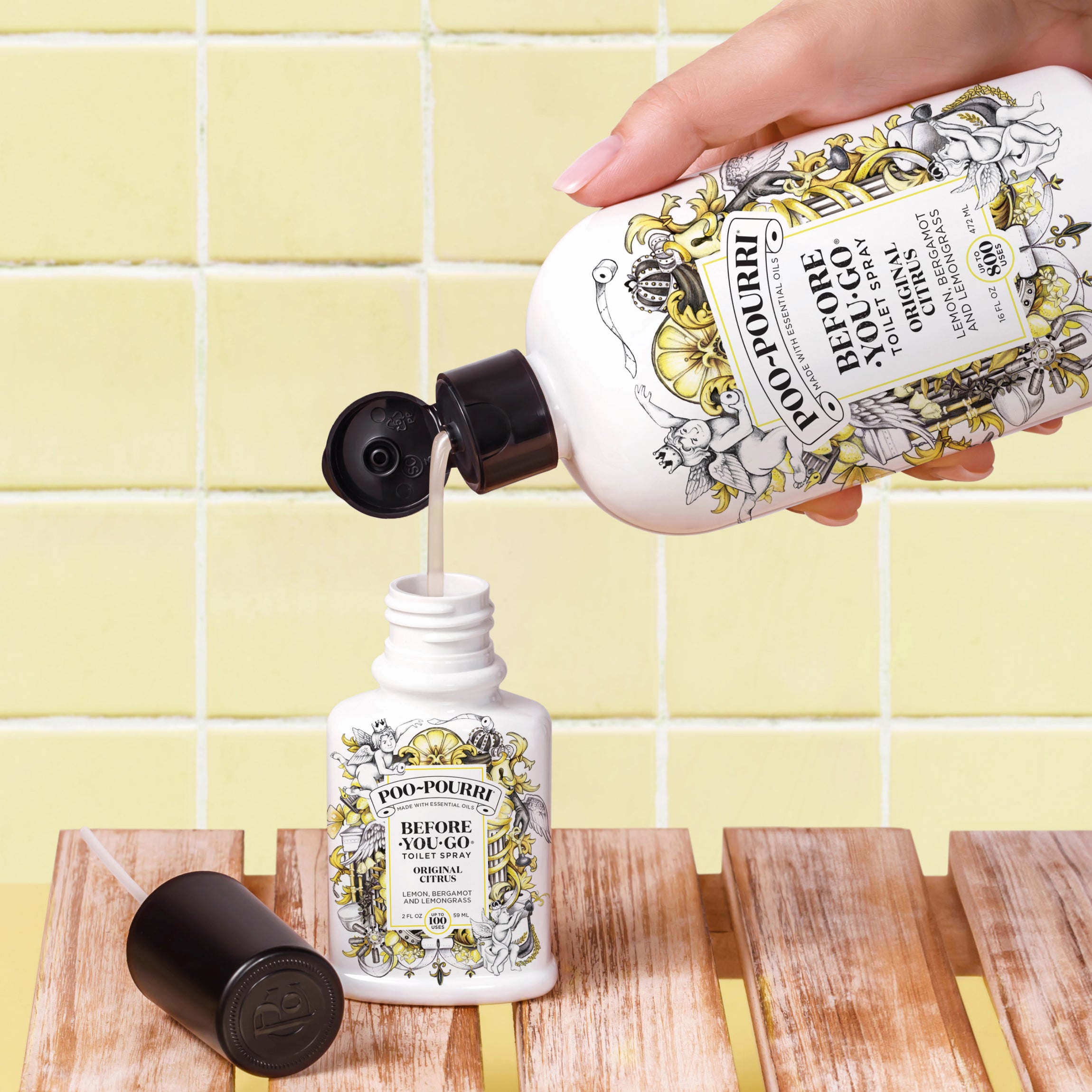 A close-up of a hand pouring Poo-Pourri “Before You Go” toilet spray from a large refill bottle into a smaller spray bottle. The bottles feature intricate black and gold designs with cherubs, and the label indicates a citrus scent. The scene is set against a yellow tiled background, with a wooden surface holding the smaller bottle and its detached cap.