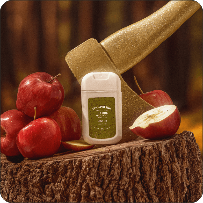 Poo~Pourro Balsam Bum toilet pocket sprayer with a green label on a wooden surface surrounded by red apples and a golden axe