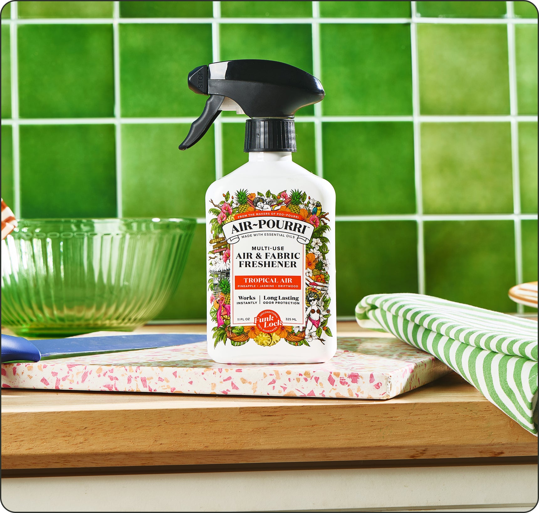 A bottle of Air~Pourri Tropical Air Air & Fabric Freshener stands on a wooden surface in front of a green tiled wall. The white spray bottle with a black trigger top is centered, with a green glass bowl on the left and a green-and-white striped cloth on the right.