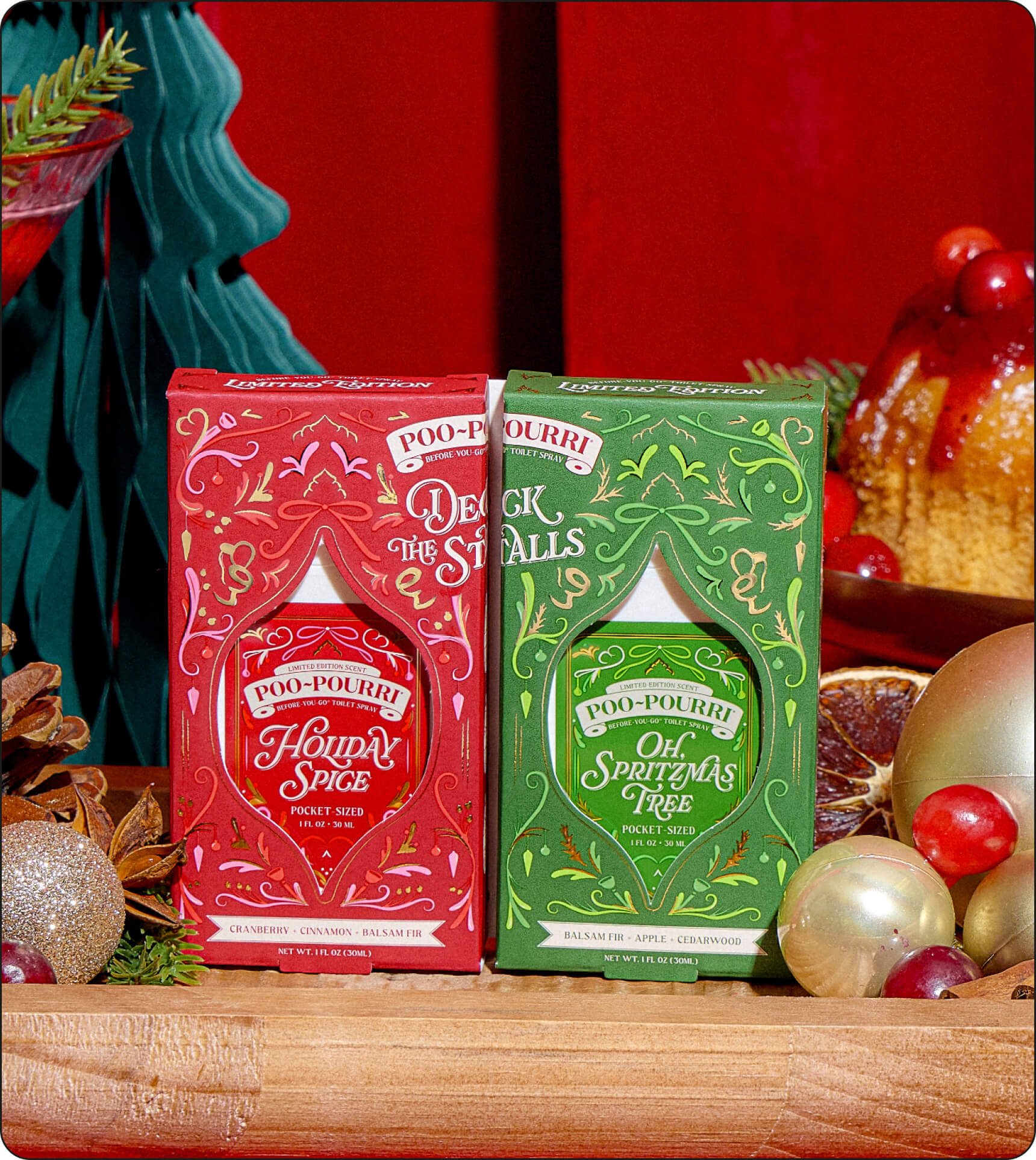 Two boxed Poo-Pourri holiday scented pocket sprayers, one red and one green, displayed on a wooden surface with Christmas decorations and a red backdrop.