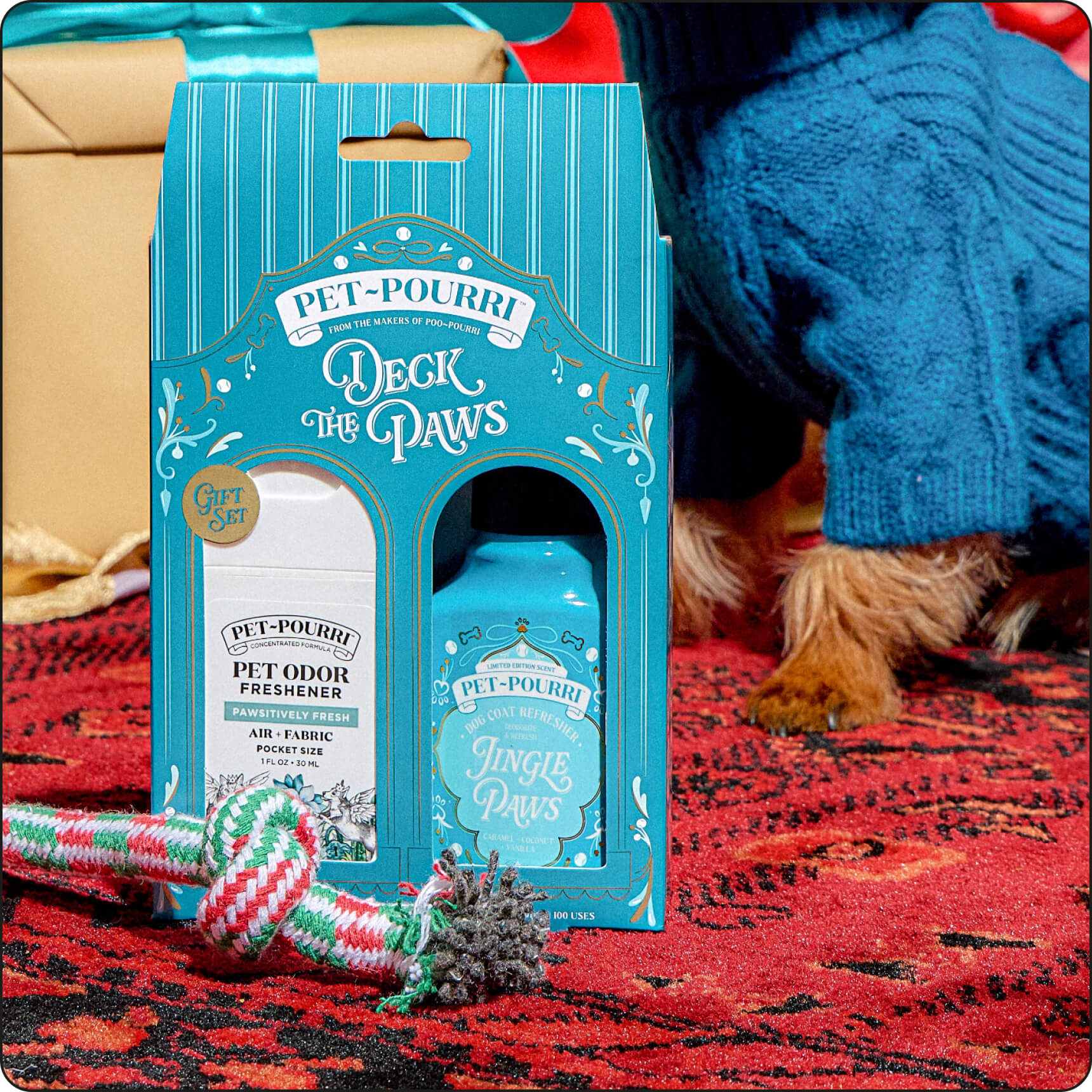 A Pet~Pourri set on a red patterned rug, with a small dog in a blue sweater visible at the right edge. The package reads Pet~Pourri and shows 'Deck the Paws' with a pet odor freshener bottle and a blue dog coat refresher spray bottle.