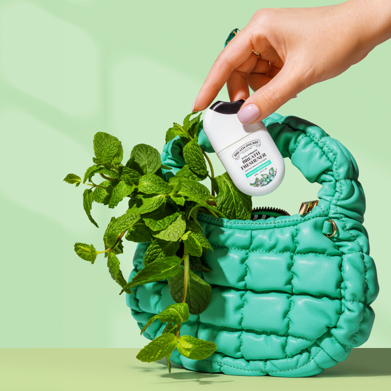 A hand places a pocket-sized Extra Strength Breath Freshener by ~Pourri into a green quilted purse, accented with mint leaves against a light green background.